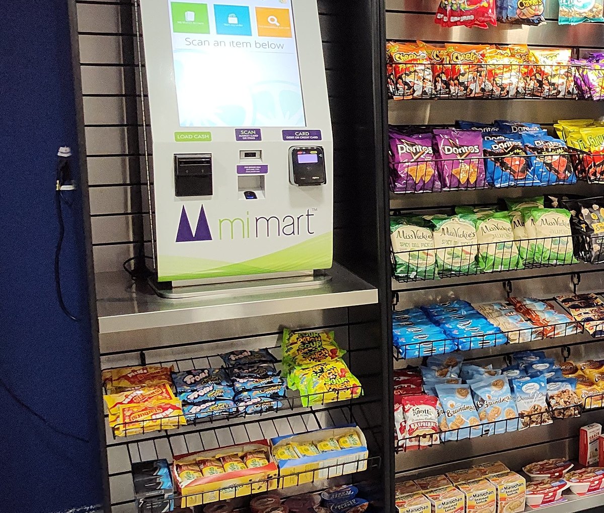 A self-checkout kiosk beside fully stocked open shelving of chips and snacks.