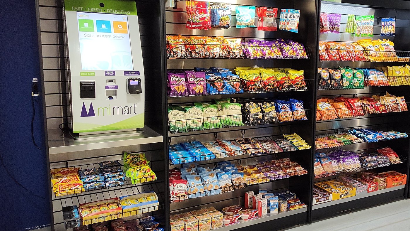An installed micro-market: a touch-screen kiosk next to long rows of shelving stocked with snacks and grab-and-go items.
