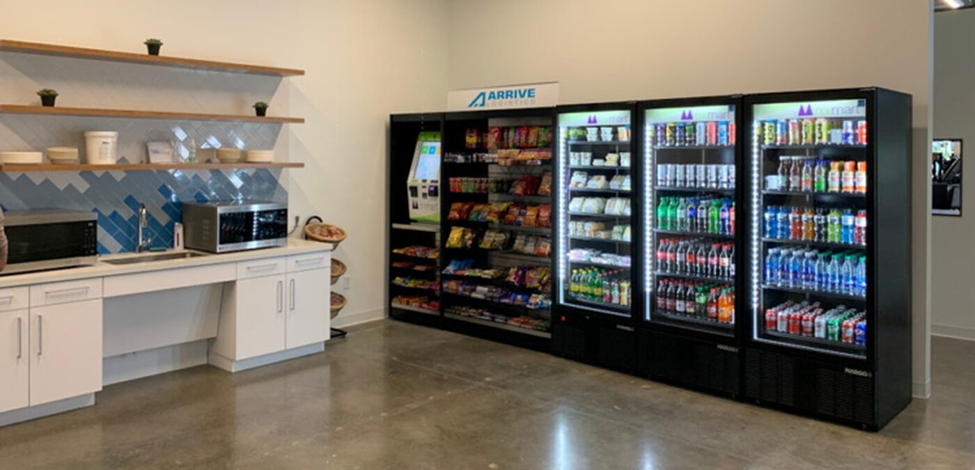 A workplace breakroom with a self-checkout micro-market, open snack shelving and three lit beverage coolers beside a kitchenette.